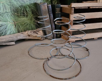 May include: A vintage metal spring with a spiral design, featuring circular base rings. The spring has a rusty, aged appearance. The background includes a wooden crate, old books, and a piece of driftwood, suggesting a rustic theme.