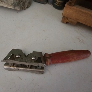 K&ouml;nnte beinhalten: Ein vintage Messer-Sch&auml;rfer aus Metall mit einem roten Holzgriff. Der Sch&auml;rfer hat zwei S&auml;tze von R&auml;dern zum Sch&auml;rfen von Messern.
