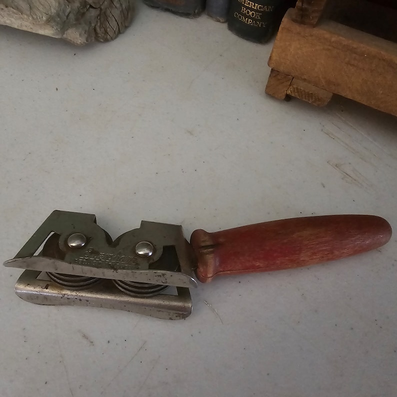 K&ouml;nnte beinhalten: Ein vintage Messer-Sch&auml;rfer aus Metall mit einem roten Holzgriff. Der Sch&auml;rfer hat zwei S&auml;tze von R&auml;dern zum Sch&auml;rfen von Messern.