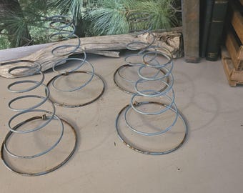 May include: Four vintage metal bed springs with a spiral design. The springs are a faded blue-gray color with some rust. The springs are on a light gray surface.