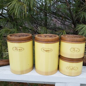 May include: A set of four cylindrical kitchen canisters with a pale yellow finish and a darker yellow base. Each canister features a brown wooden lid with a carved floral pattern and a decorative oval label with the text Flour, Sugar, Tea, or Coffee.
