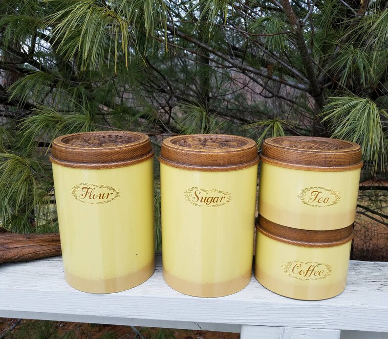 May include: A set of four cylindrical kitchen canisters with a pale yellow finish and a darker yellow base. Each canister features a brown wooden lid with a carved floral pattern and a decorative oval label with the text Flour, Sugar, Tea, or Coffee.