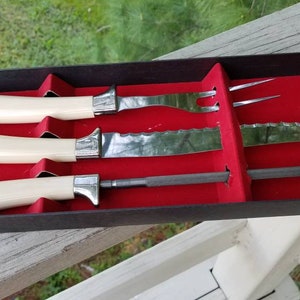 Puede incluir: Un juego de cuatro cuchillos de cocina con mangos blancos y hojas plateadas en una caja forrada de terciopelo rojo. El juego incluye un cuchillo para trinchar, un cuchillo para pan, un cuchillo para carne y un tenedor.