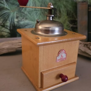 May include: A vintage wooden coffee grinder with a metal top and handle. The grinder has a small drawer and a red knob. A red, teardrop-shaped handle is attached to the top. The grinder has a label that reads "TRUSSER".