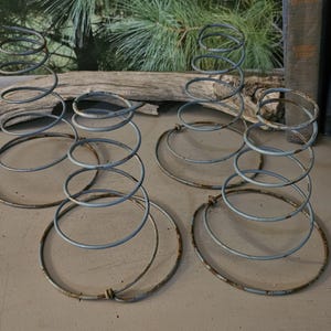 May include: Four vintage metal bed springs with a spiral design. The springs are silver and have a circular base. The background includes a piece of driftwood, books, and a wooden crate.