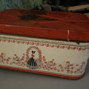 May include: Vintage red and white metal cookie tin with a floral design and a woman standing under an archway. The lid has a rusty patina.