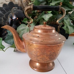 May include: A copper teapot with an ornate floral design and a black wooden handle. The teapot has a round body and a spout on the side.
