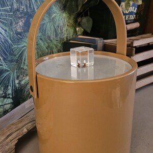 May include: A vintage, mustard-colored ice bucket with a matching handle. The cylindrical bucket has a clear lid and a clear, square handle on top. The background includes greenery and a wooden surface.