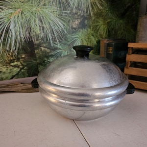 May include: A vintage aluminum cooking pot with a lid and black handle. The pot has a textured surface and a black knob on top. The pot is round with a double-banded design around the middle. The pot is sitting on a white surface.