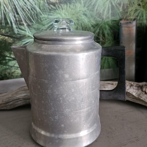 May include: A brushed aluminum percolator coffee pot featuring a cylindrical body, a wide spout, and a black handle. The pot includes a matching aluminum lid topped with a small, clear glass knob. The exterior surface shows a matte metallic finish.