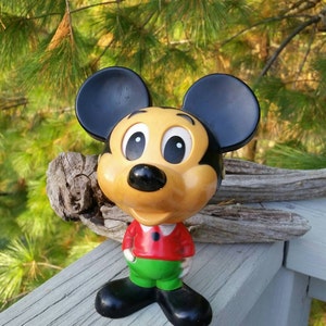 May include: A vintage plastic Mickey Mouse figurine with black ears, large eyes, a yellow face, a red shirt, and green pants.