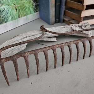 May include: An antique, rusty metal garden rake with a curved handle and multiple tines. The rake is lying on a gray surface, with a piece of driftwood in the background. The rake's design suggests it is for gardening.