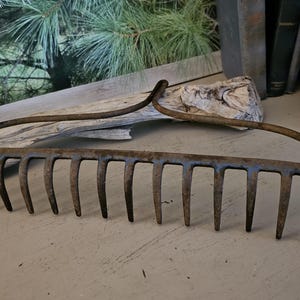 May include: An antique metal garden rake with a curved handle and multiple tines. The rake is weathered with a rusty brown patina, suggesting age and use. The tool is designed for gardening and landscaping.