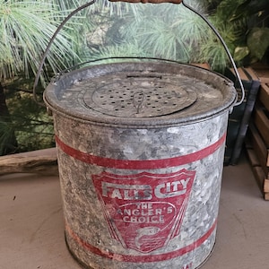 May include: Vintage galvanized metal bait bucket with a wooden handle. The bucket has a red and white graphic that reads "FALLS CITY THE ANGLER'S CHOICE". The bucket has a perforated lid and a wire handle.