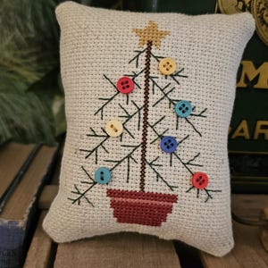 May include: A small, square, off-white cross-stitch pillow featuring a Christmas tree design. The tree is brown with colorful button ornaments and a gold star. The tree sits in a red pot. The pillow is on a wooden surface.