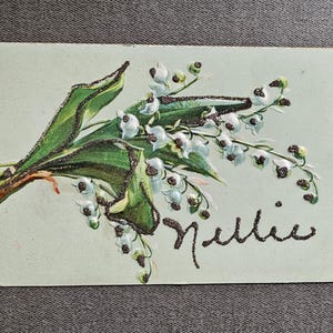 May include: Vintage postcard featuring a detailed illustration of lily of the valley flowers with green leaves and white bell-shaped blooms. The word "Nellie" is written in a glittery script. The postcard has a light green background.