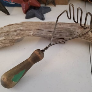 May include: A vintage metal potato masher with a green and brown wooden handle. The masher has a twisted wire handle and a curved, multi-pronged head.