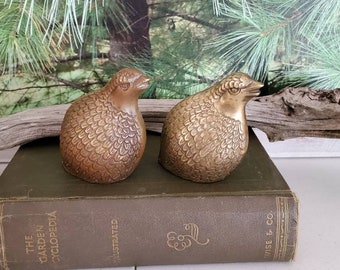 May include: Two gold-colored metal bird figurines with detailed feather patterns. The birds are facing each other and are sitting on a brown book.