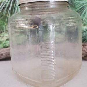 May include: A clear glass jar with a white lid. The jar has a square shape and a textured surface.