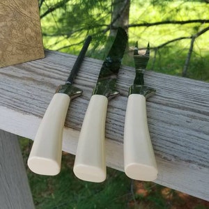 Puede incluir: Tres cuchillos de cocina con mangos blancos y hojas plateadas. Los cuchillos est&aacute;n sobre una superficie de madera.