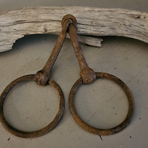 May include: A pair of rusted iron rings connected by two metal bars that meet at a single top loop. The metal has a dark, weathered, and oxidized brown texture throughout the entire structure, showing signs of age and exposure to the elements.