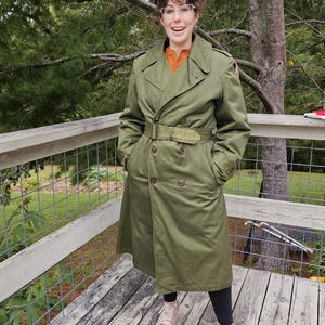 May include: An olive green double-breasted trench coat featuring a matching waist belt, wide lapels, and shoulder epaulets. The coat is worn over an orange collared shirt, black leggings, and beige sneakers, with a small circular patch on the left shoulder.