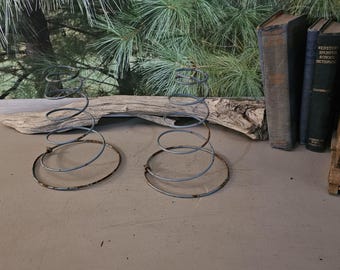 May include: Two antique metal bed springs with a spiral design. The springs are silver and brown, with a circular base. The background includes old books, a wooden crate, and a piece of driftwood.
