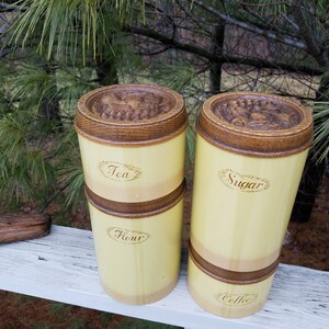 May include: A set of four cylindrical kitchen canisters with a pale yellow finish and dark brown wooden accents. The canisters feature embossed lids and labels reading Tea, Flour, Sugar, and Coffee in a cursive script font on the front surfaces.