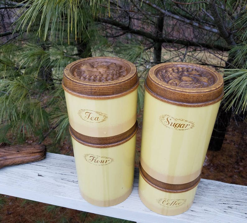 May include: A set of four cylindrical kitchen canisters with a pale yellow finish and dark brown wooden accents. The canisters feature embossed lids and labels reading Tea, Flour, Sugar, and Coffee in a cursive script font on the front surfaces.