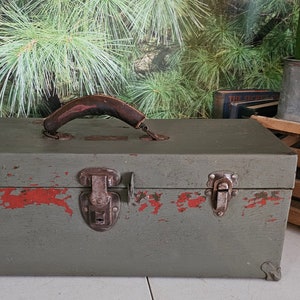 May include: A vintage green metal toolbox with a wooden handle and rusty metal latches. The toolbox has red paint chipping off the surface.