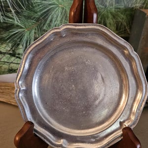 May include: A round, silver-toned metal plate with a scalloped, wavy edge and a smooth, slightly weathered surface. The plate features a raised rim and a flat center, displaying a metallic sheen with subtle signs of age and texture throughout.