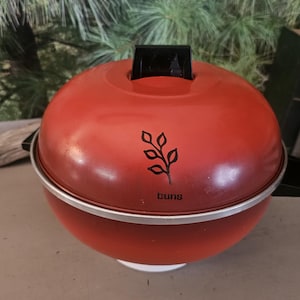 May include: A red, spherical, vintage-style barbecue with a black handle on top. The barbecue has a silver band around the middle and a white base. The word "buns" is printed on the front, below a leaf design.
