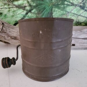 May include: A vintage metal flour sifter with a crank handle. The sifter is brown and has the words "ONE CUP" printed on the side.