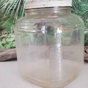 May include: A clear glass jar with a white lid. The jar has a square shape and a textured surface.