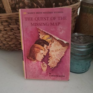 May include: A pink Nancy Drew mystery book titled "The Quest of the Missing Map" by Carolyn Keene. The book cover features an illustration of three girls looking at a map.