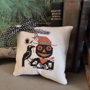 May include: A white pillow with a cross-stitch design of a scarecrow with a black crow. The scarecrow is wearing a brown hat and has a pumpkin face. The pillow is decorated with a black and white polka dot ribbon.