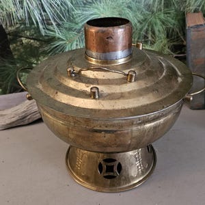 May include: Vintage brass hot pot with a copper chimney. The pot has a round base with decorative cutouts and a lid with a handle. The pot is designed for cooking and serving food. The pot is made of brass and copper.