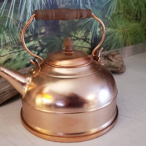 May include: A copper tea kettle with a wooden handle. The kettle has a shiny, polished finish and a rounded body.