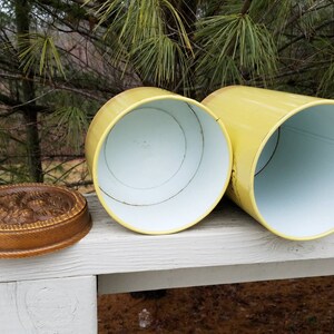 May include: Two cylindrical yellow metal canisters with white interiors and separate circular wooden lids featuring a carved floral pattern on the top surface. The canisters are positioned horizontally with their open ends facing forward.