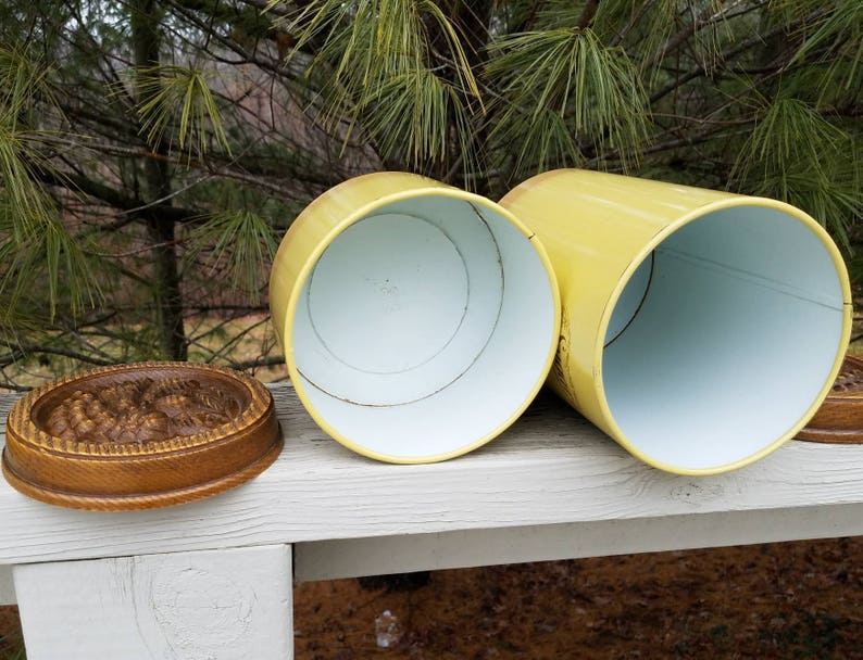 May include: Two cylindrical yellow metal canisters with white interiors and separate circular wooden lids featuring a carved floral pattern on the top surface. The canisters are positioned horizontally with their open ends facing forward.