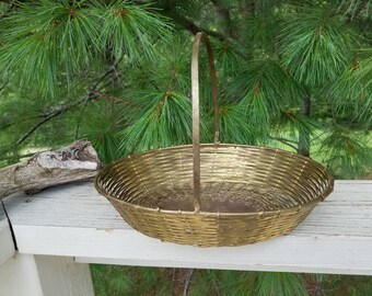 May include: A gold-toned woven basket with a metal handle. The basket is round and has a woven pattern. The basket is sitting on a white surface.