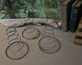 May include: Three vintage metal bed springs with a spiral design. The springs are a faded blue color and sit on a neutral surface. The background includes old books, a wooden crate, and a natural wood element.