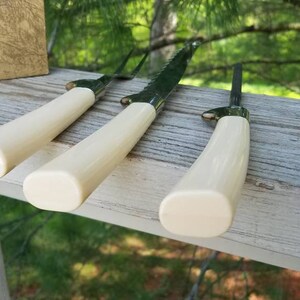 Puede incluir: Tres cuchillos con mangos de color marfil y hojas plateadas. Los cuchillos est&aacute;n dispuestos sobre una superficie de madera.