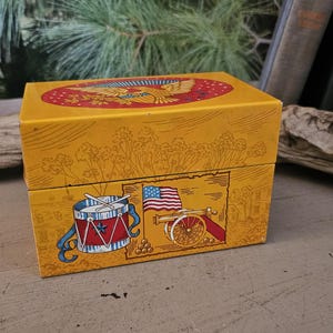 May include: A rectangular yellow box with illustrations of a drum, American flag, and cannon. The top of the box features a red oval with a golden eagle. The box is likely vintage or antique.