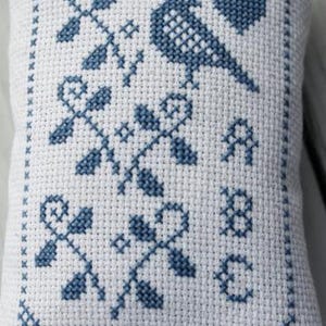 May include: A small, white, cross-stitched pillow with a blue bird, heart, and floral design. The pillow has a blue border and the letters "A", "8", and "E" stitched on it. The design is symmetrical and detailed.