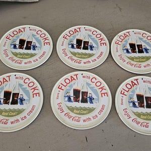 Könnte beinhalten: Sechs Vintage Coca-Cola Untersetzer. Jeder runde Untersetzer hat einen weißen Hintergrund und den Text "FLOAT with COKE" in Rot und Weiß. Die Untersetzer haben ein Retro-Design mit einer Coca-Cola-Flasche und Eiscreme.