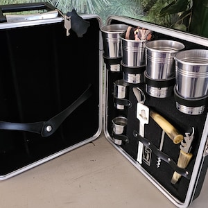 May include: Open vintage black and silver bar kit with various stainless steel cups, a key, and bar tools. The interior is lined with black fabric. The kit is open, revealing the contents, and has a handle for carrying.