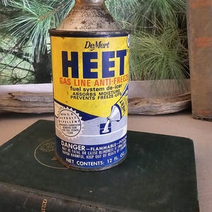 May include: Vintage DeMert HEET gas line anti-freeze can with yellow and blue label. The can, with a metal cap, sits on a dark green book. The label states the product absorbs moisture and prevents freeze-ups. The can contains 12 fl oz.