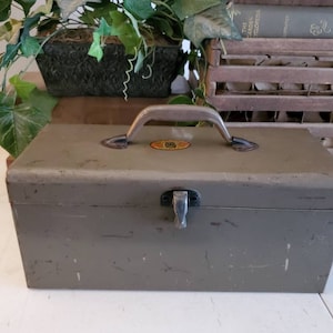 May include: A vintage metal toolbox with a silver handle and a worn green paint finish. The toolbox has a latch on the front and a small metal plate with a logo on the top.