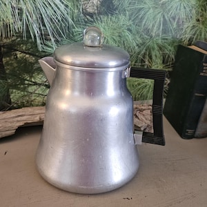May include: A vintage aluminum coffee percolator with a black handle and a clear glass knob on the lid. The percolator is silver and sits on a table. Behind it are two books and a wooden crate.